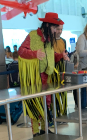 Person in a red hat and yellow fringed outfit stands by a railing at an indoor event.