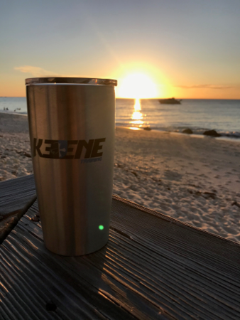 Metal tumbler on a deck at sunset, with calm ocean and beach in the background.