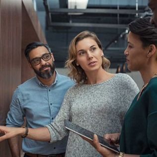Three coworkers talking in an office hallway, one holding a tablet.