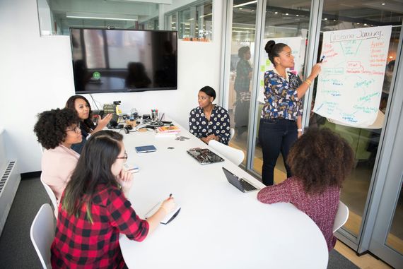 Team meeting in a glass-walled conference room, with one person presenting at a whiteboard.