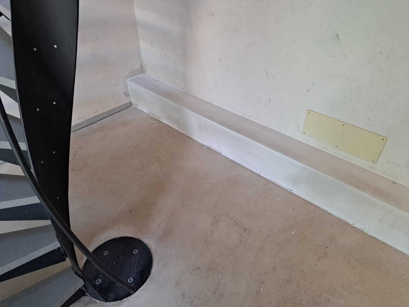 Scala a chiocciola con ringhiera nera accanto a una parete bianca con battiscopa e una piccola apertura.