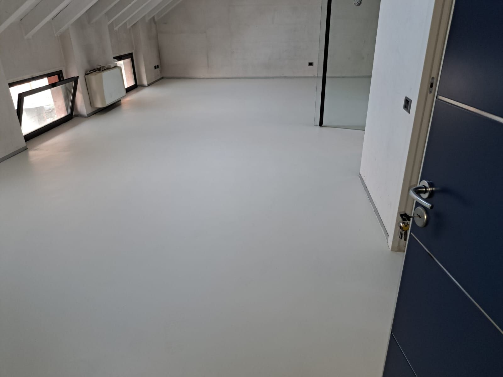 Stanza vuota con pavimento grigio chiaro, soffitto spiovente, porta e finestre blu scuro.