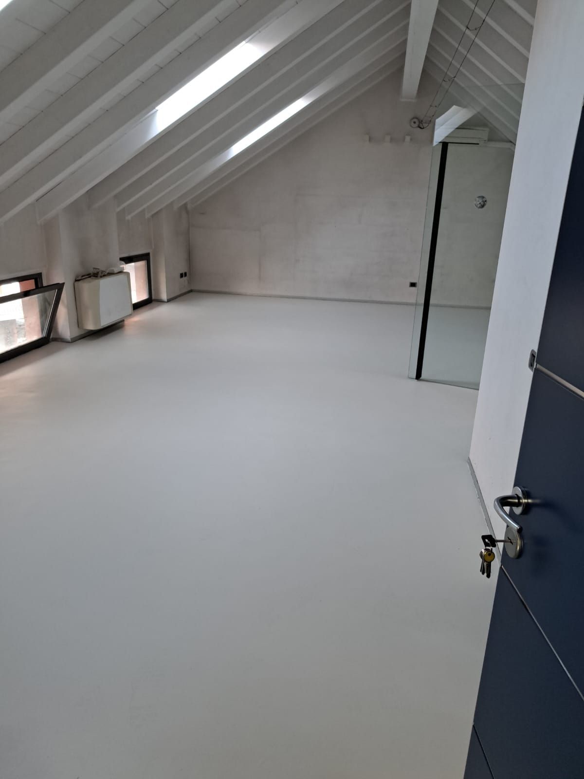 Mansarda vuota con pareti, pavimento e soffitto bianchi, lucernari e una porta blu scuro.
