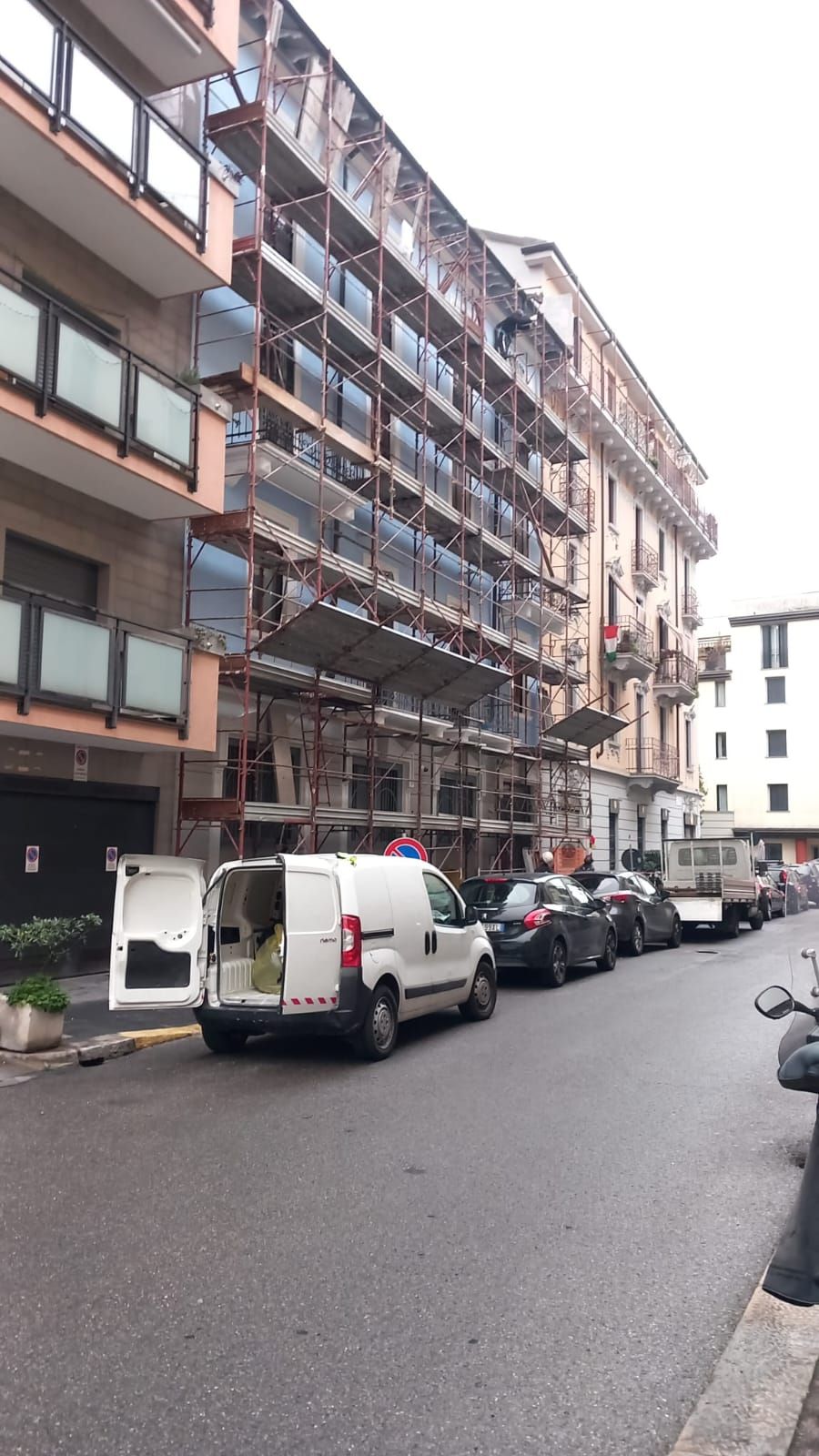 Un'impalcatura ricopre la facciata di un edificio, un furgone bianco è parcheggiato sulla strada di fronte, le auto sono parcheggiate lungo il marciapiede.