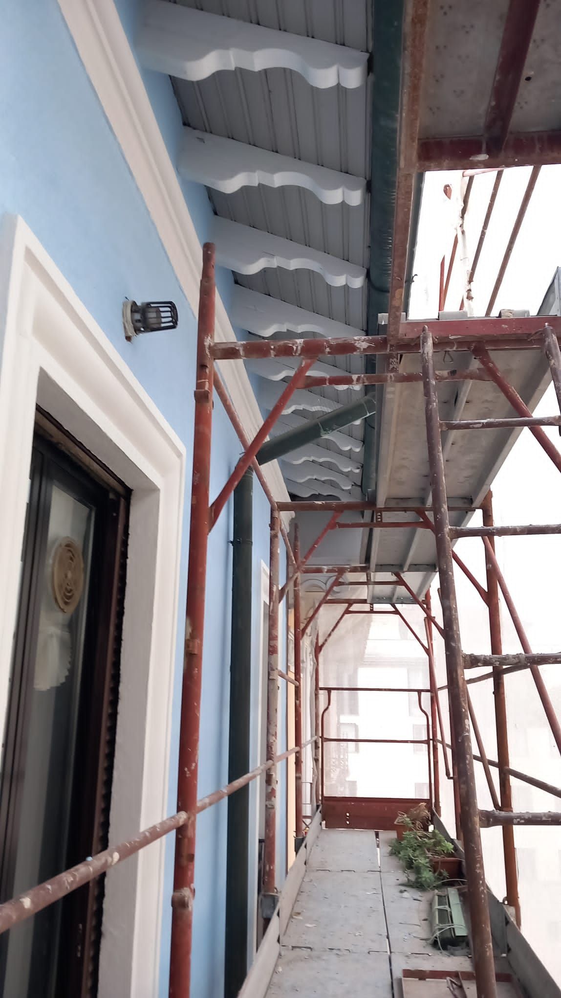 Un'impalcatura sullo sfondo di un edificio azzurro con finiture bianche; una persona sta lavorando sul tetto in legno.