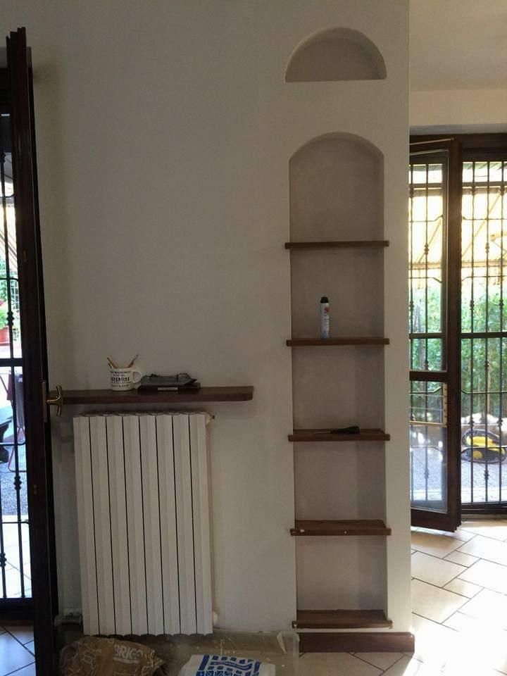Parete interna con mensole integrate, termosifone e due porte in vetro. Parete bianca, mensole in legno, stipiti delle porte scuri.