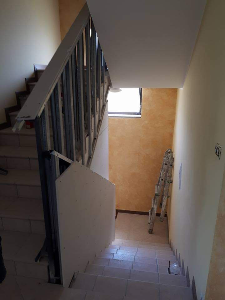 Scala con corrimano e ascensore chiuso. La scala poggia contro una parete arancione testurizzata.