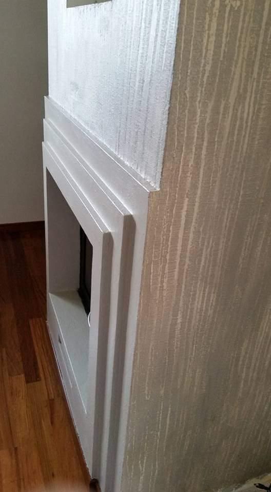 Caminetto bianco con cornice a strati, contro una parete strutturata e un pavimento in legno.