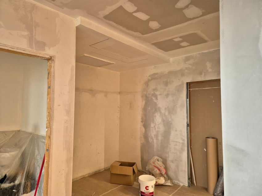 Stanza in costruzione: pareti beige, controsoffitto, porta aperta, scatola di cartone, secchio di vernice.