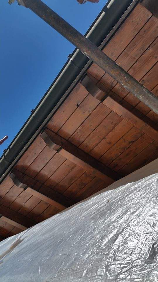 Travi di legno del tetto sotto un cielo azzurro, sostenute da impalcature e ricoperte di plastica.
