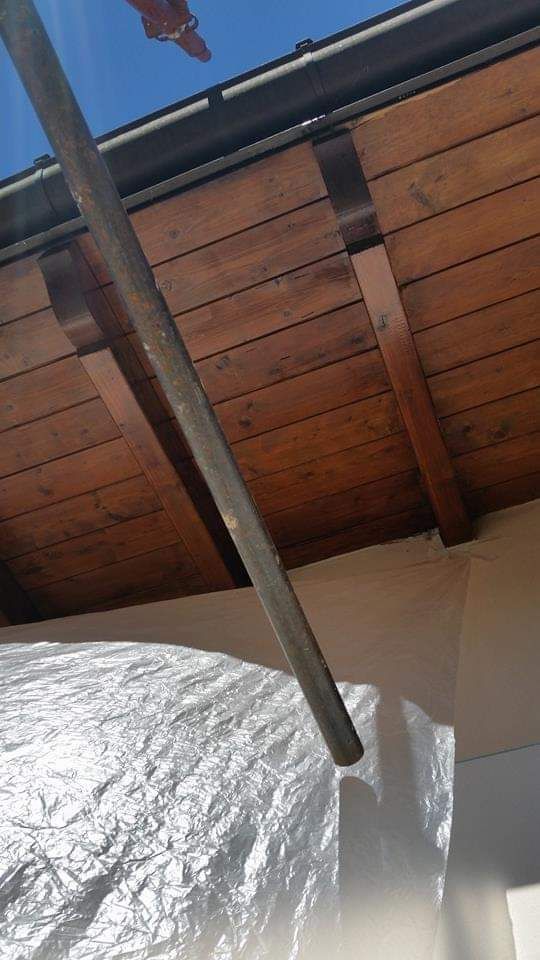 Soffitto in legno marrone con travi, tubo grigio inclinato verso il basso, telo bianco sottostante, cielo azzurro visibile.