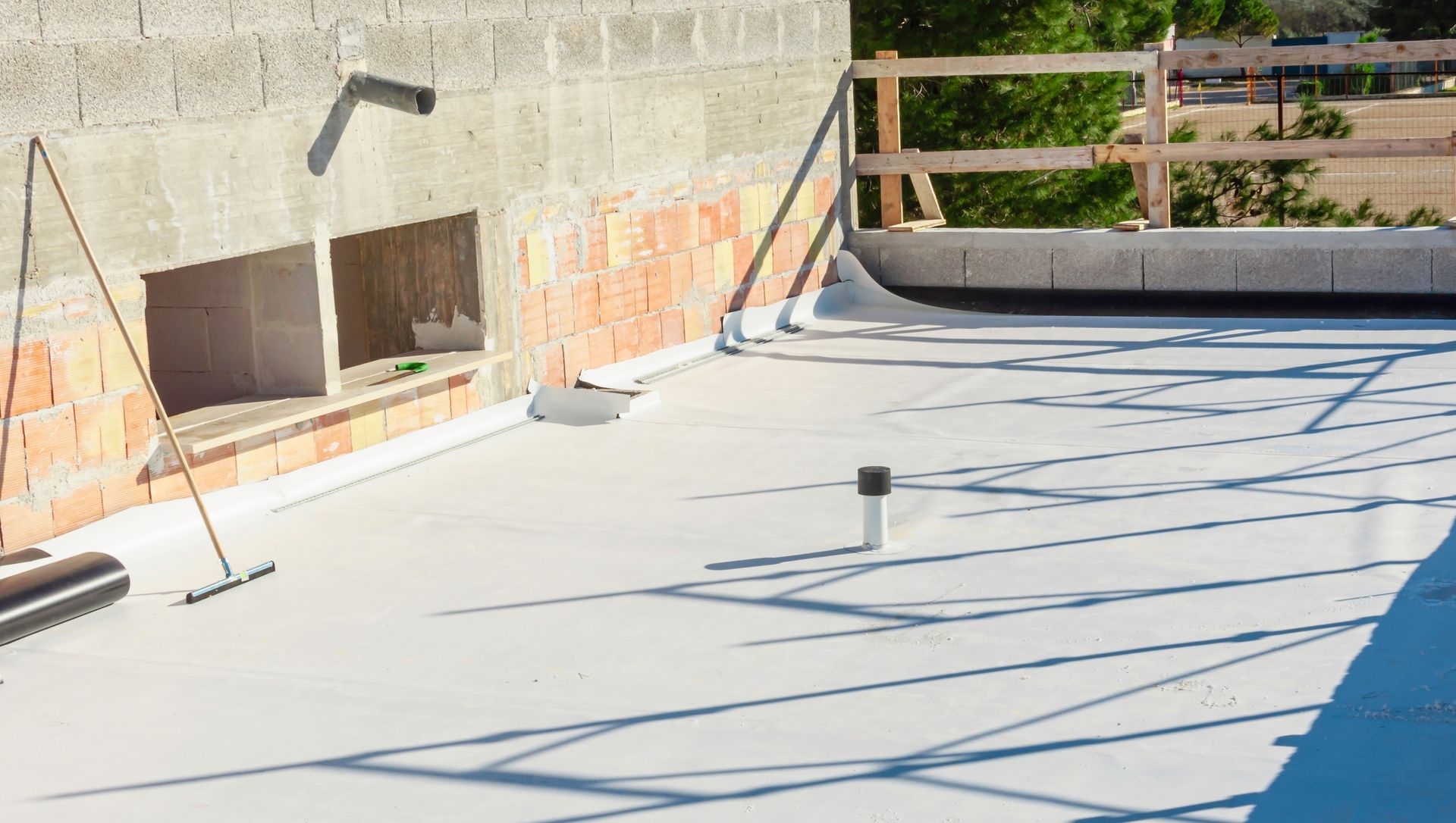 Flat rooftop with light-colored surface, drainage pipes, brick wall section, and wooden safety railing.