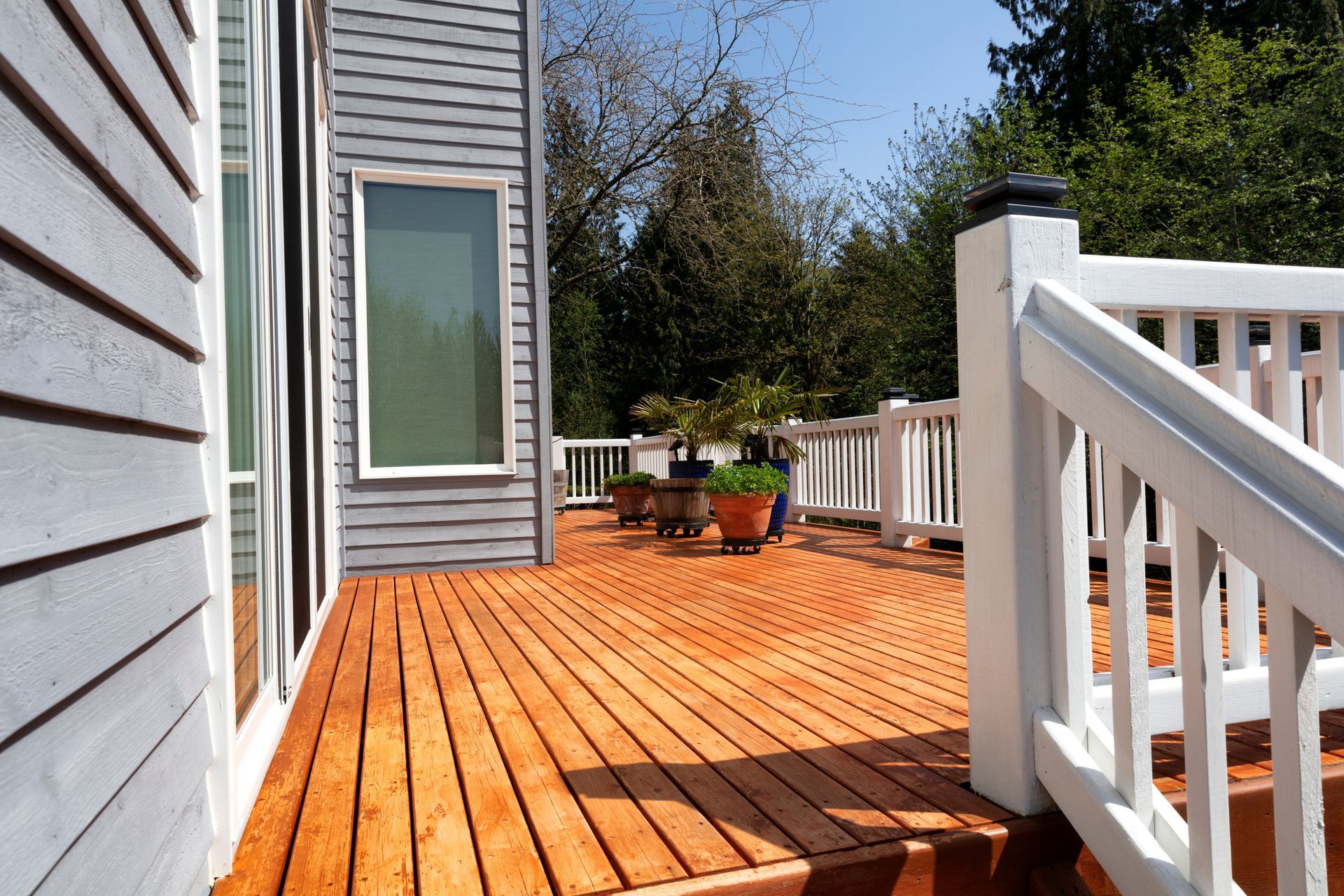 Wooden deck with white railing next to a gray house, potted plants in the background.