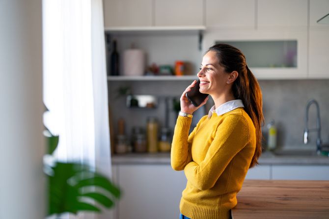 Woman in yellow sweater smiles while talking on the phone in a kitchen.