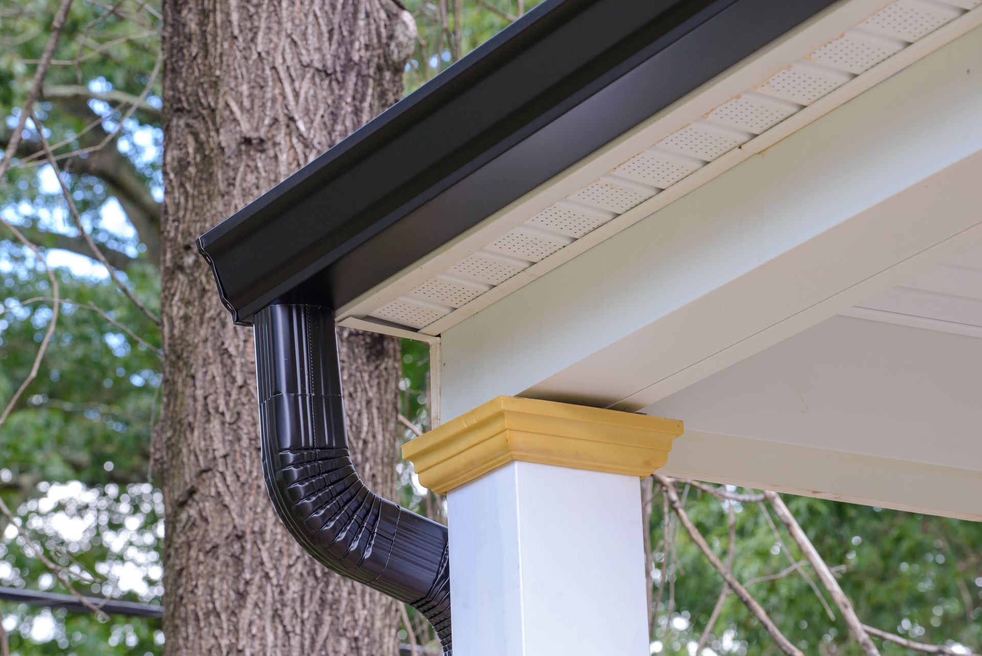 Black gutter and downspout on a white porch column with a yellow top against a tree and foliage.