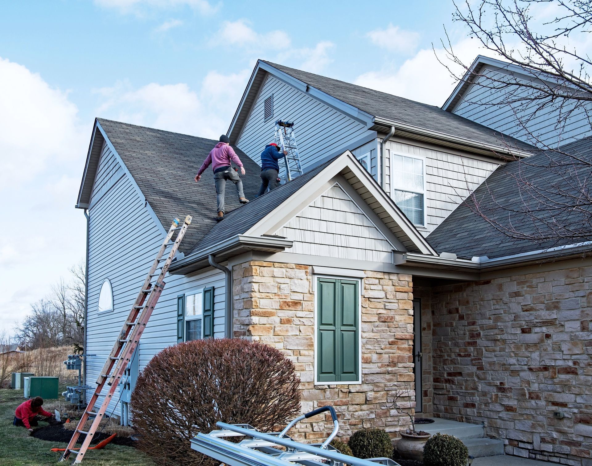 Two workers on a roof, installing shingles, with a ladder and tools. House has stone siding and blue siding.