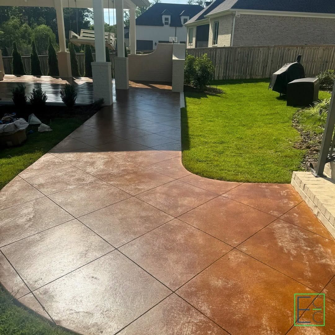 Sunlit stamped concrete patio leading to a green backyard with fence, shrubs, and an outdoor chair.