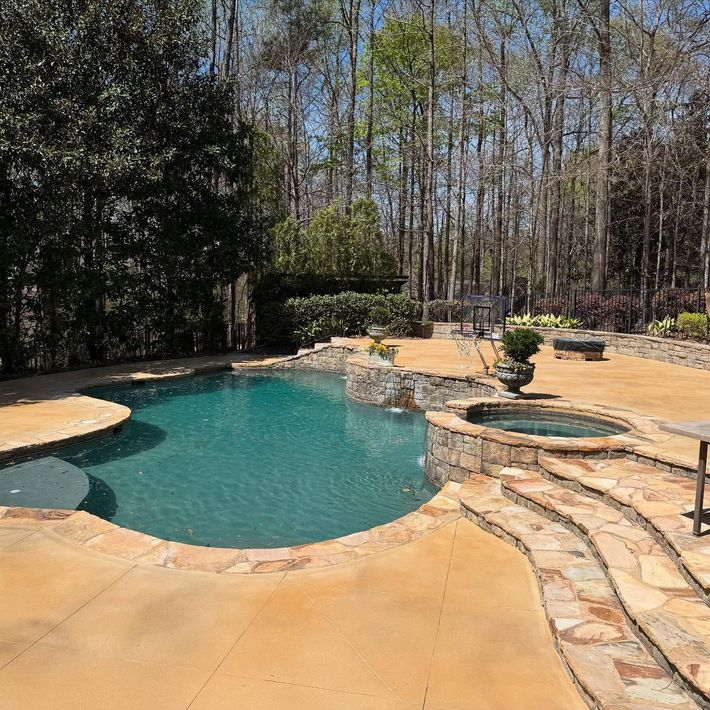Backyard pool with stone patio, hot tub, and tall trees in the background