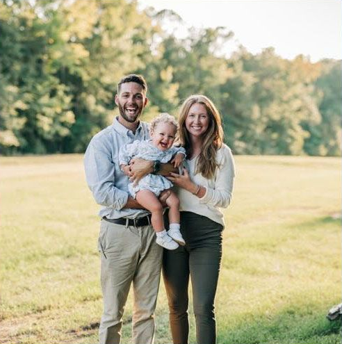 Smiling family of three standing outdoors in a sunlit park, holding a baby.
