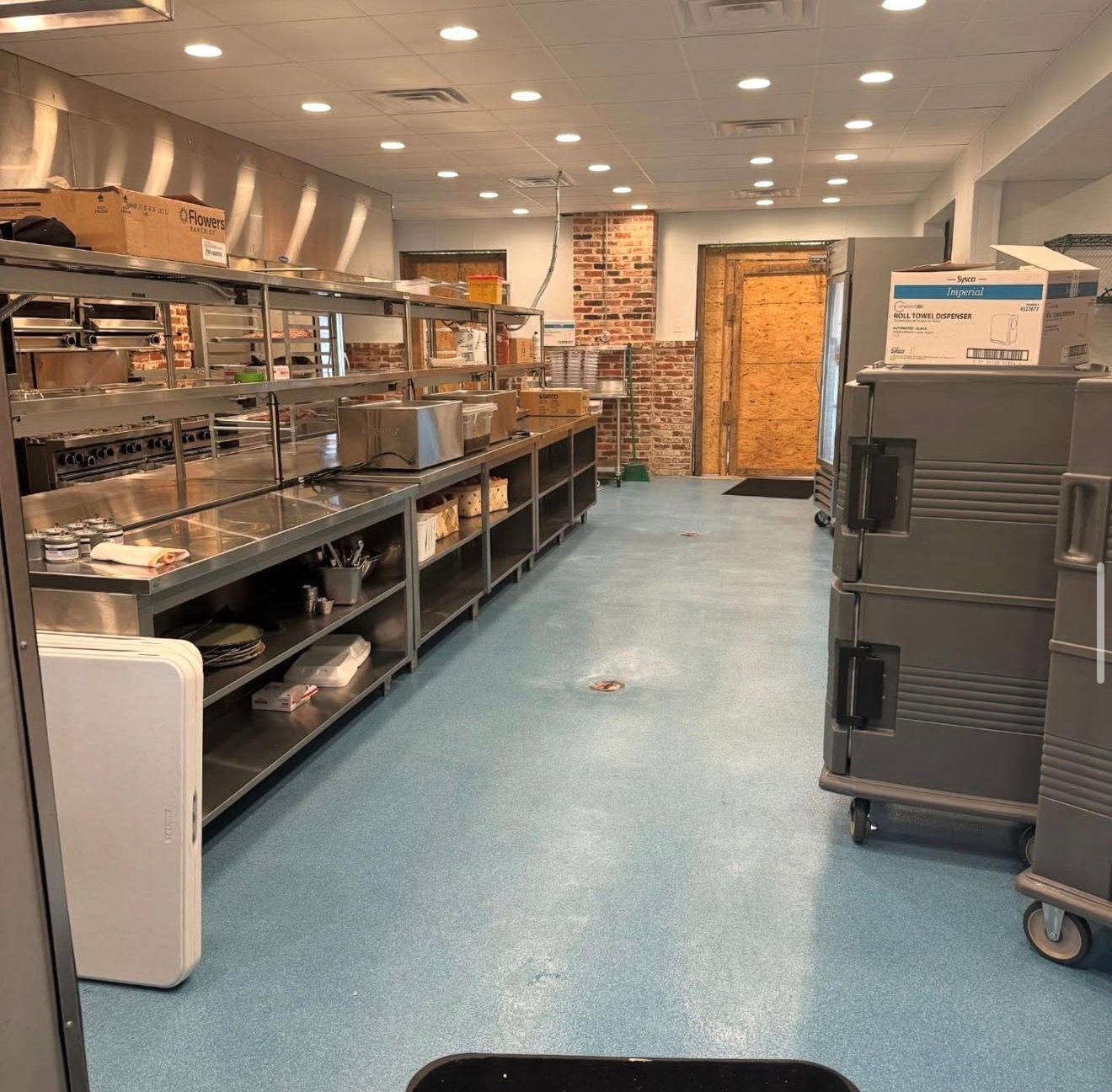 Commercial kitchen with metal shelving, stacked racks, blue floor, and cardboard boxes near a back door