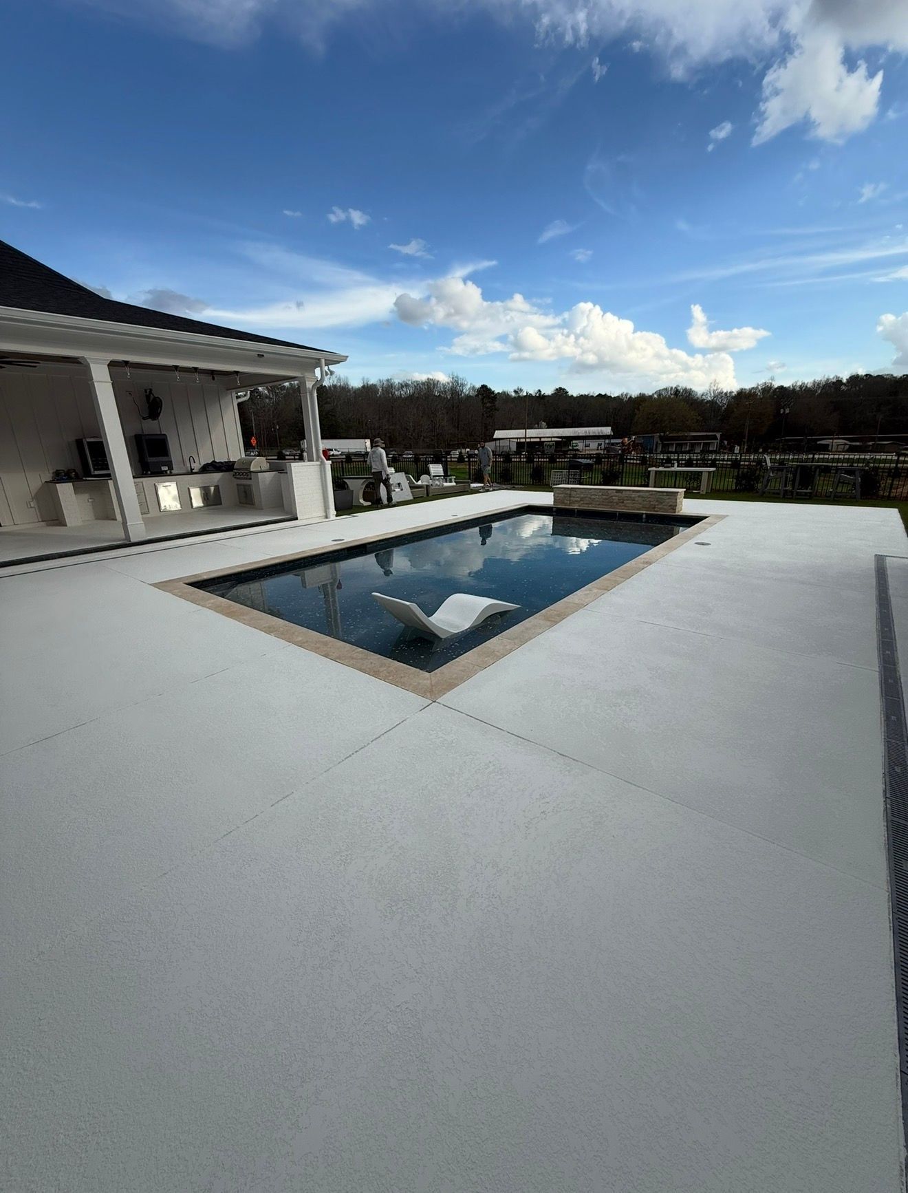 Modern patio with a rectangular pool, white decking, and a house under a blue sky
