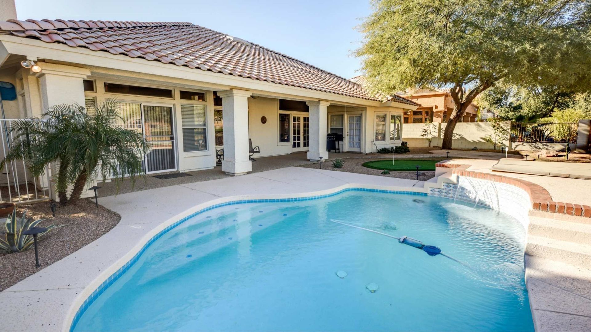 Backyard with swimming pool, patio, and home exterior. Blue water, white trim, beige tile. Sunny setting.