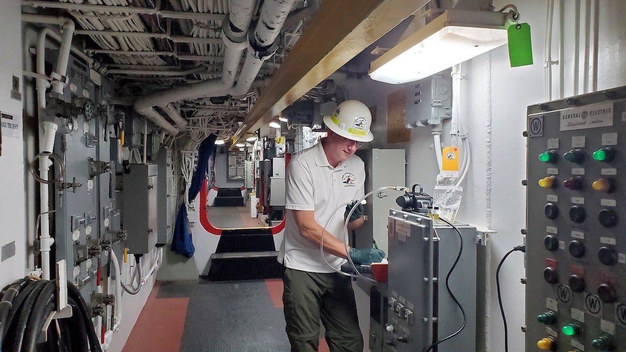 Worker in hard hat checking equipment in a narrow industrial corridor with pipes and control panels