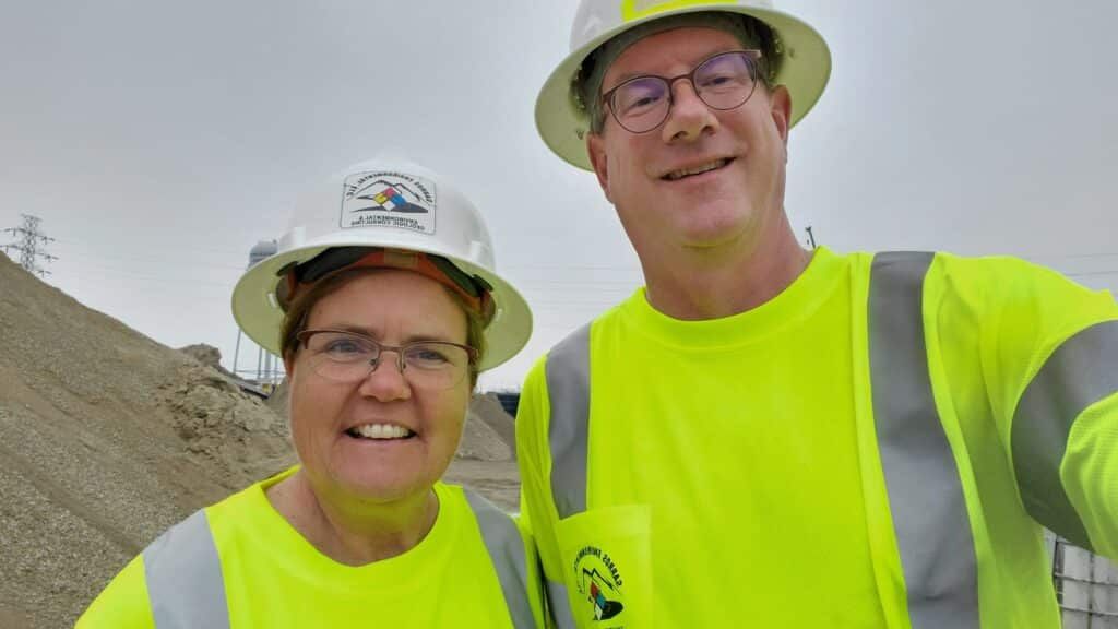 Two people in neon yellow safety shirts and hard hats posing outdoors at a worksite