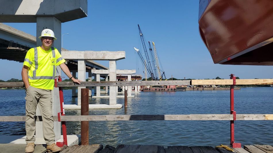 Worker in neon safety vest standing by a waterfront railing with cranes and water in the background