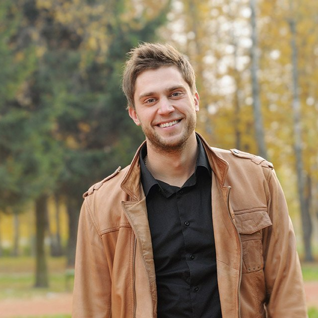 Smiling man wearing a brown leather jacket and black shirt, standing outdoors with trees in the background.