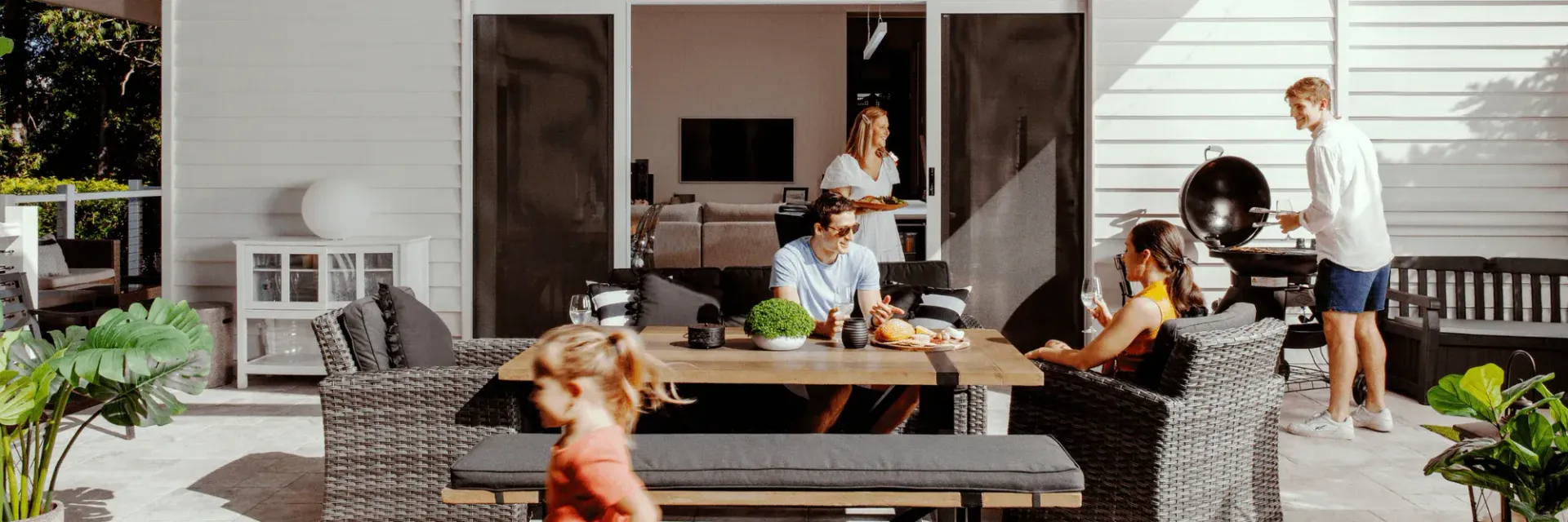 Family gathered on a patio for a barbecue. People are seated around a table and grilling.