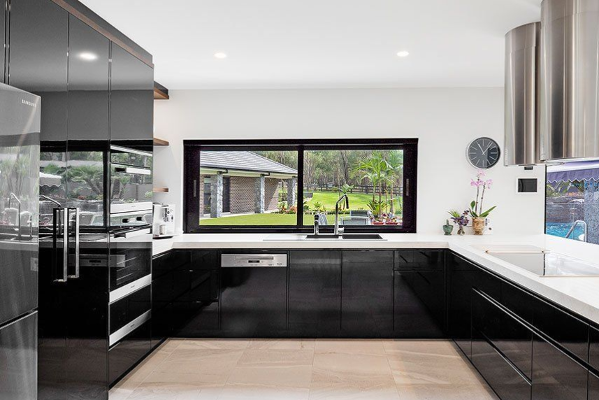 Modern kitchen with black glossy cabinets, white countertops, large window overlooking a yard.