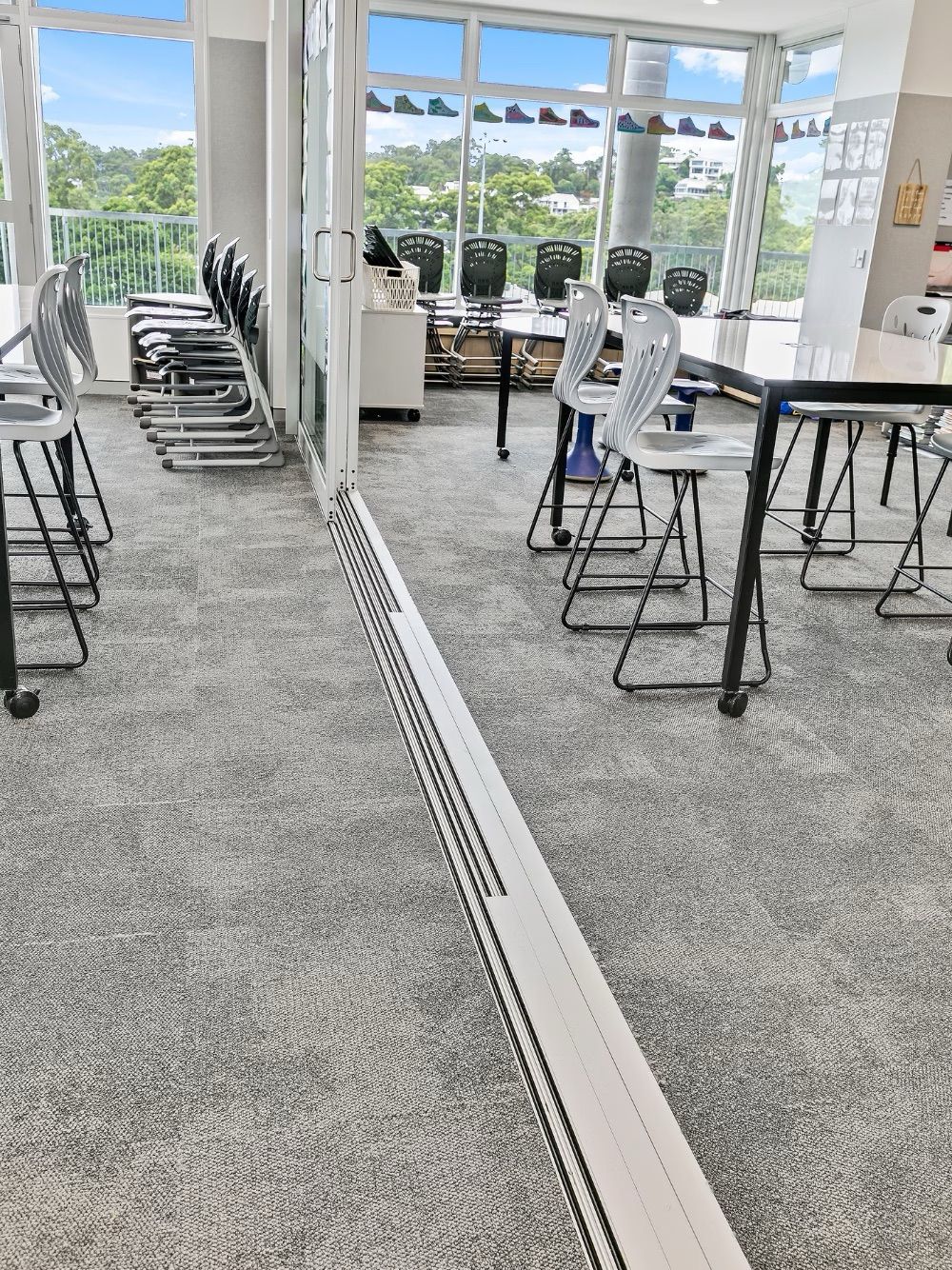 Classroom with desks, chairs, and computers. Windows overlook trees and blue sky. Grey carpeted floor.