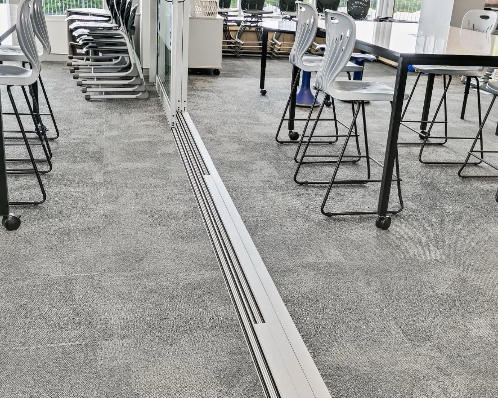 Gray carpeted classroom with glass wall track and high chairs.