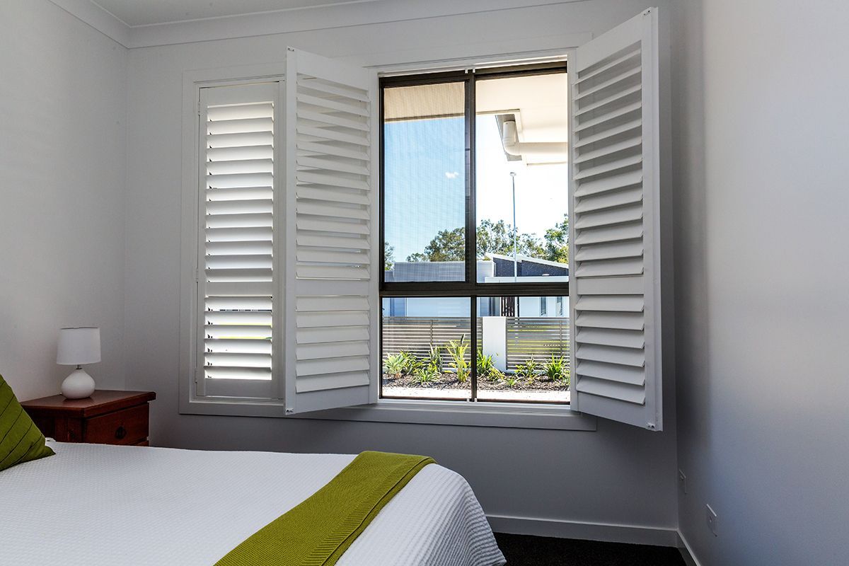 Bedroom window with white shutters open, revealing a view of a sunny outdoor area.
