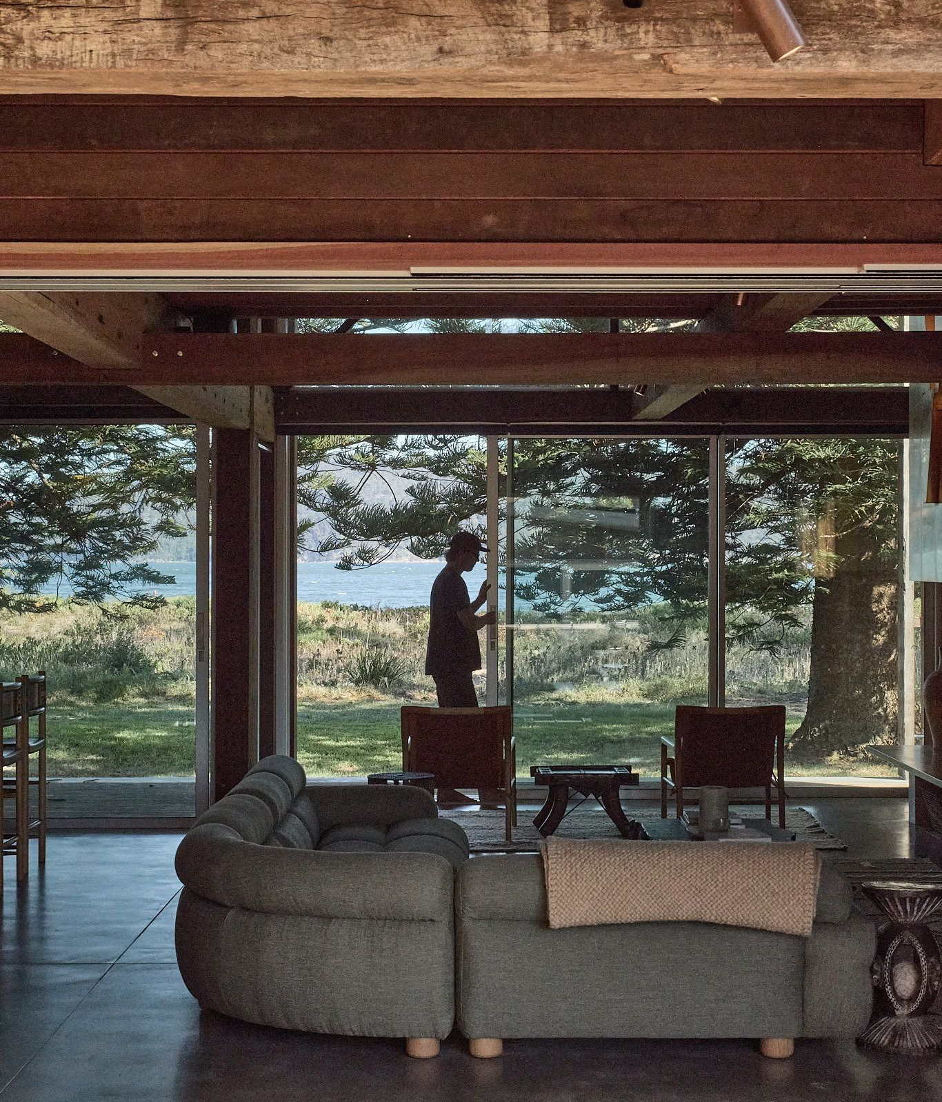 Living room with glass walls, wood beams, and a view of a person standing outside by trees and water.