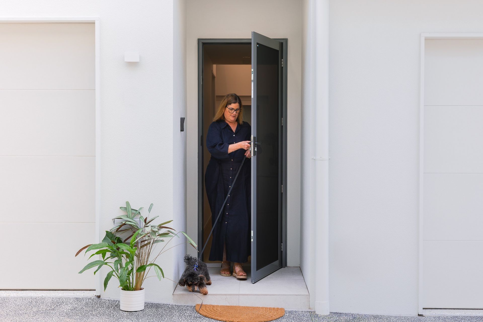 Woman opening a dark gray screen door. A small dog is at her feet. White house exterior.