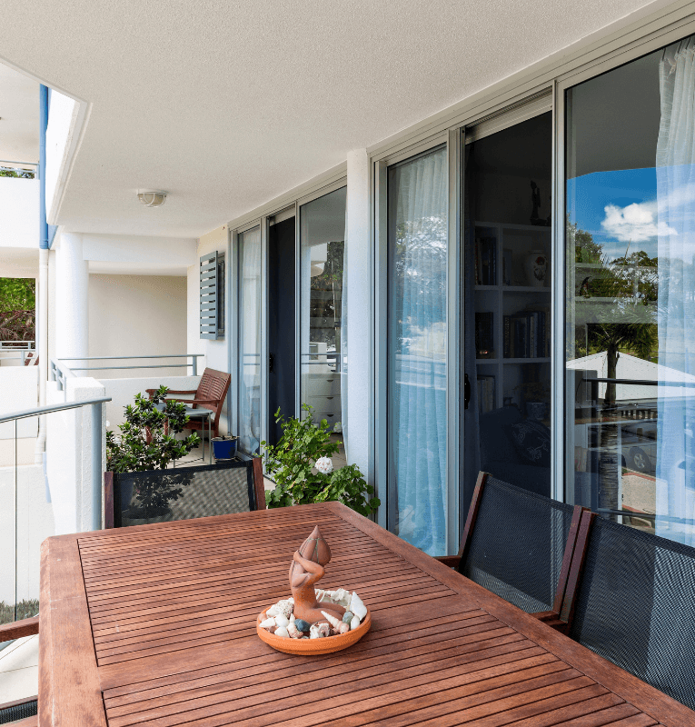 Balcony with wooden table, chairs, plants, and glass sliding doors with white curtains.