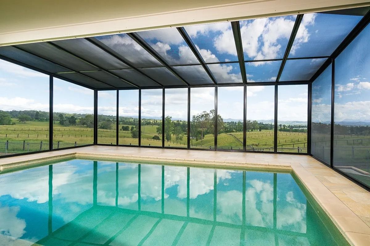 Indoor swimming pool with clear glass walls and ceiling, overlooking a green landscape and blue sky with clouds.