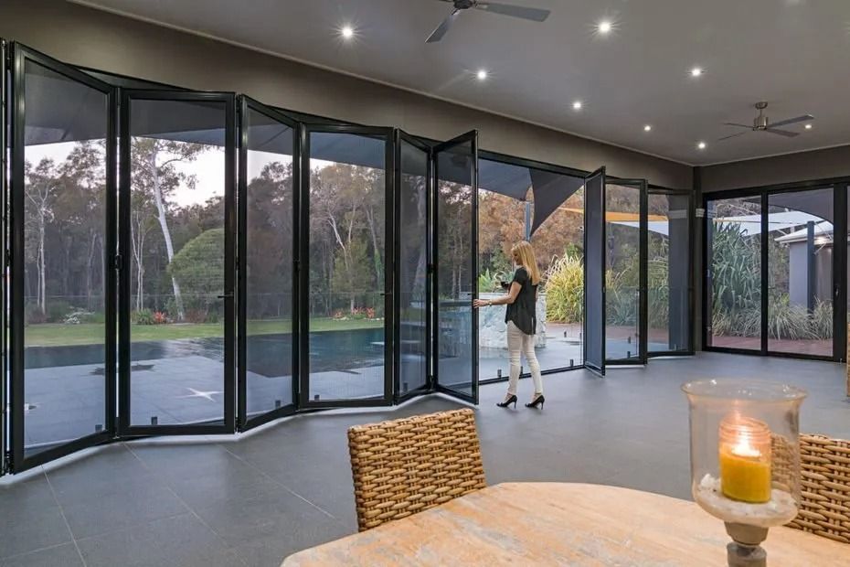 A woman opens folding glass doors to a backyard pool area. The room has gray flooring and a neutral color palette.