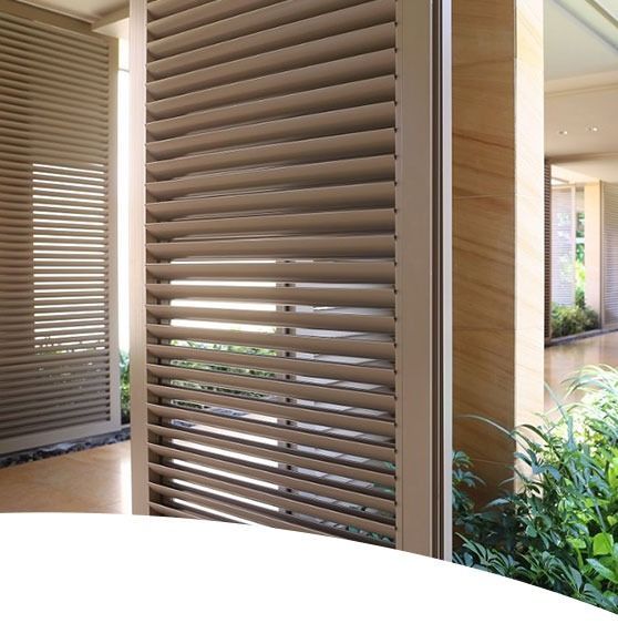 Beige louvered shutters, providing privacy, separate an outdoor hallway with sunlight and plants.