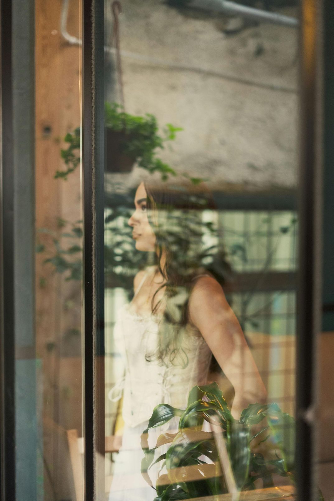 Woman in white corset stands by a window, looking out. Green plants and brick exterior are visible.