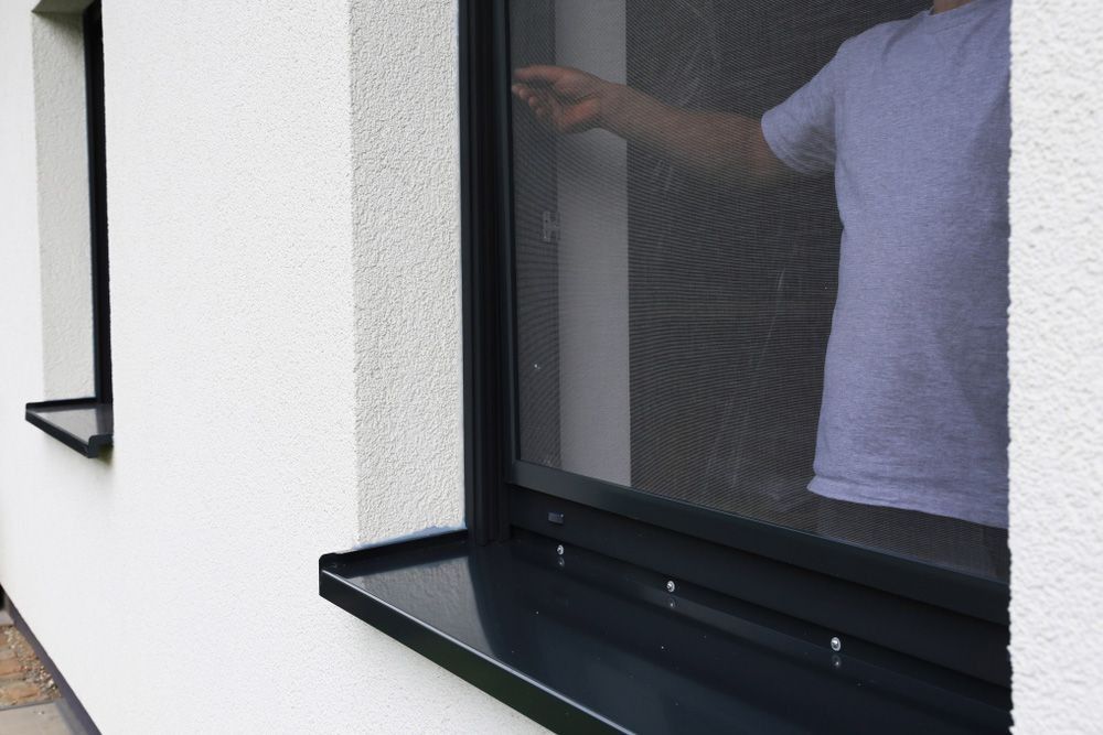 Person's hand reaching toward a black window screen on a white wall.