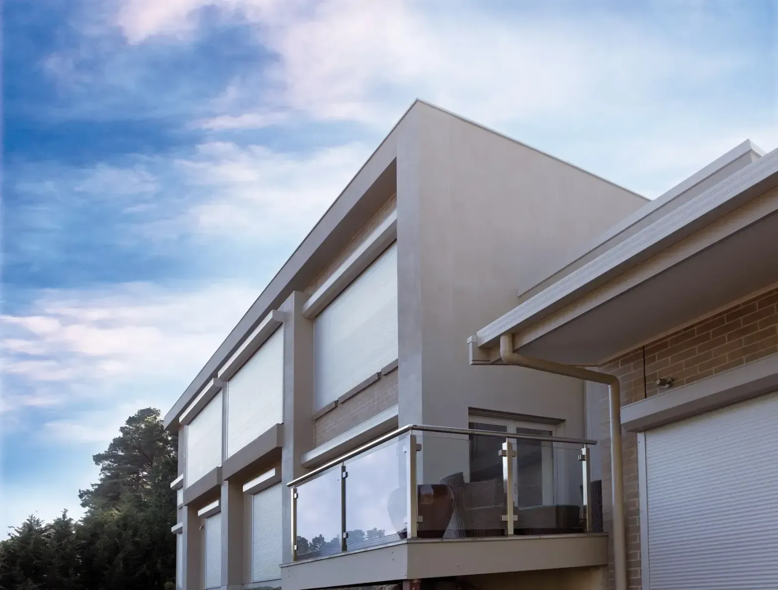 Modern beige house with balcony, glass railing, and outdoor blinds. Bright blue sky with clouds in the background.