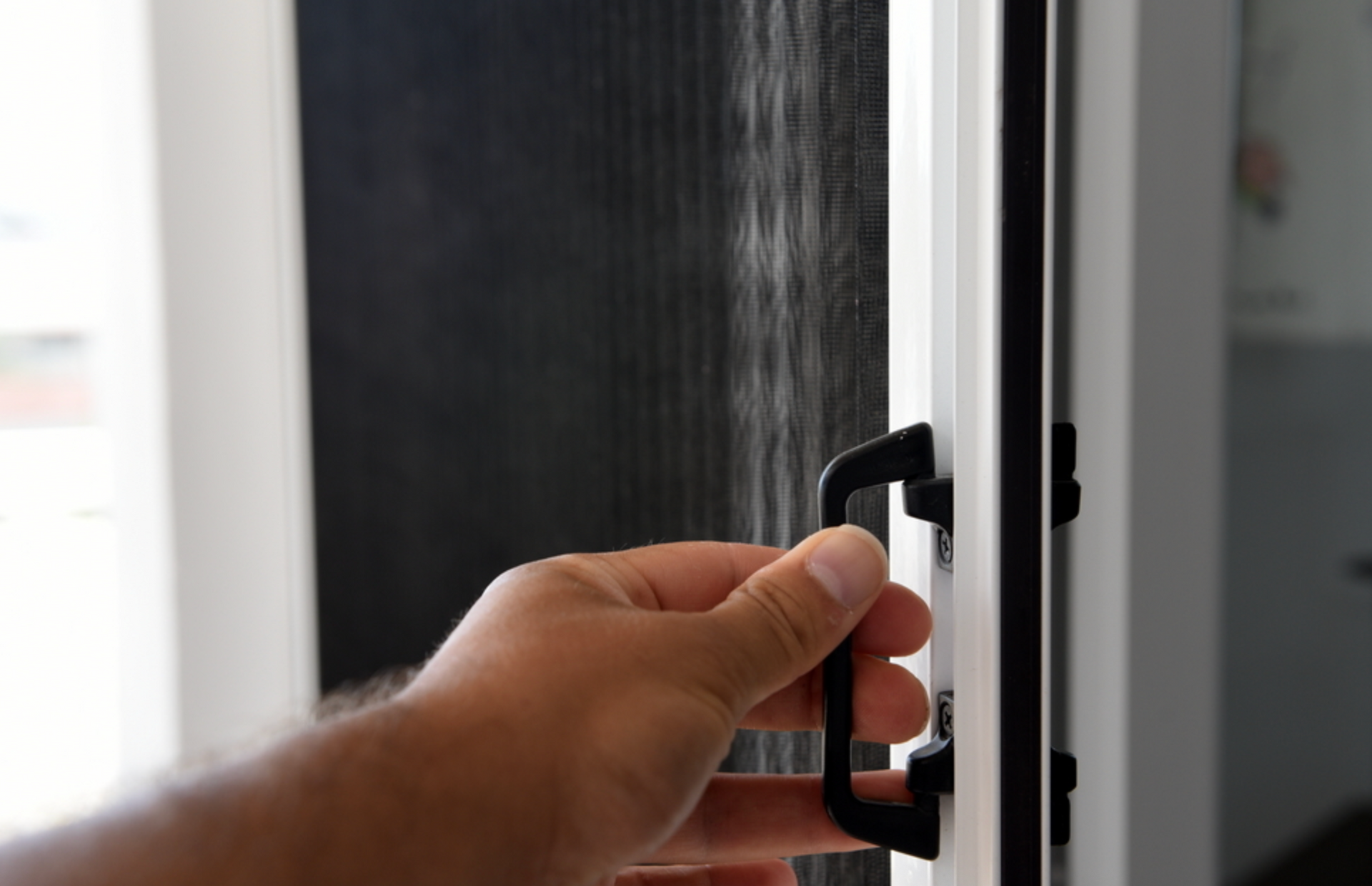 Hand pulls a black door handle on a white frame, opening a screen door.