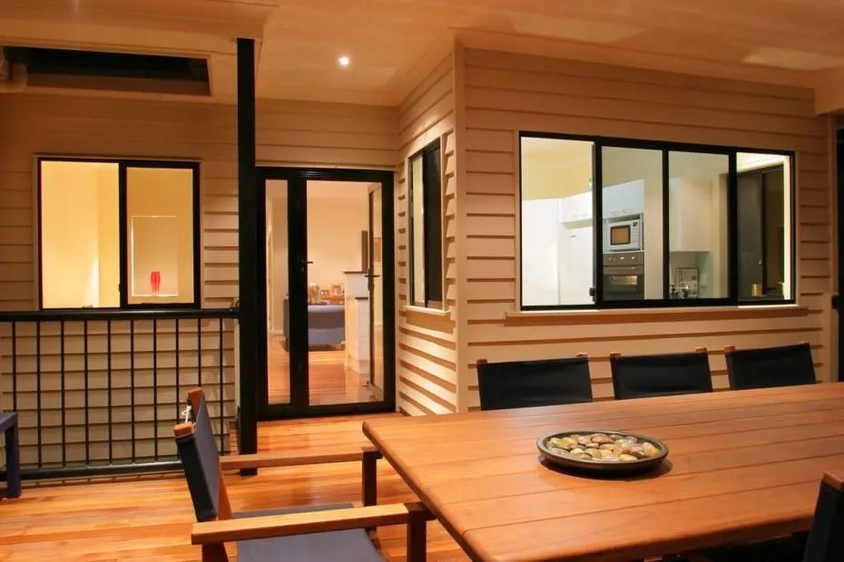 Outdoor dining area with wooden table, chairs, and windows; interior visible.