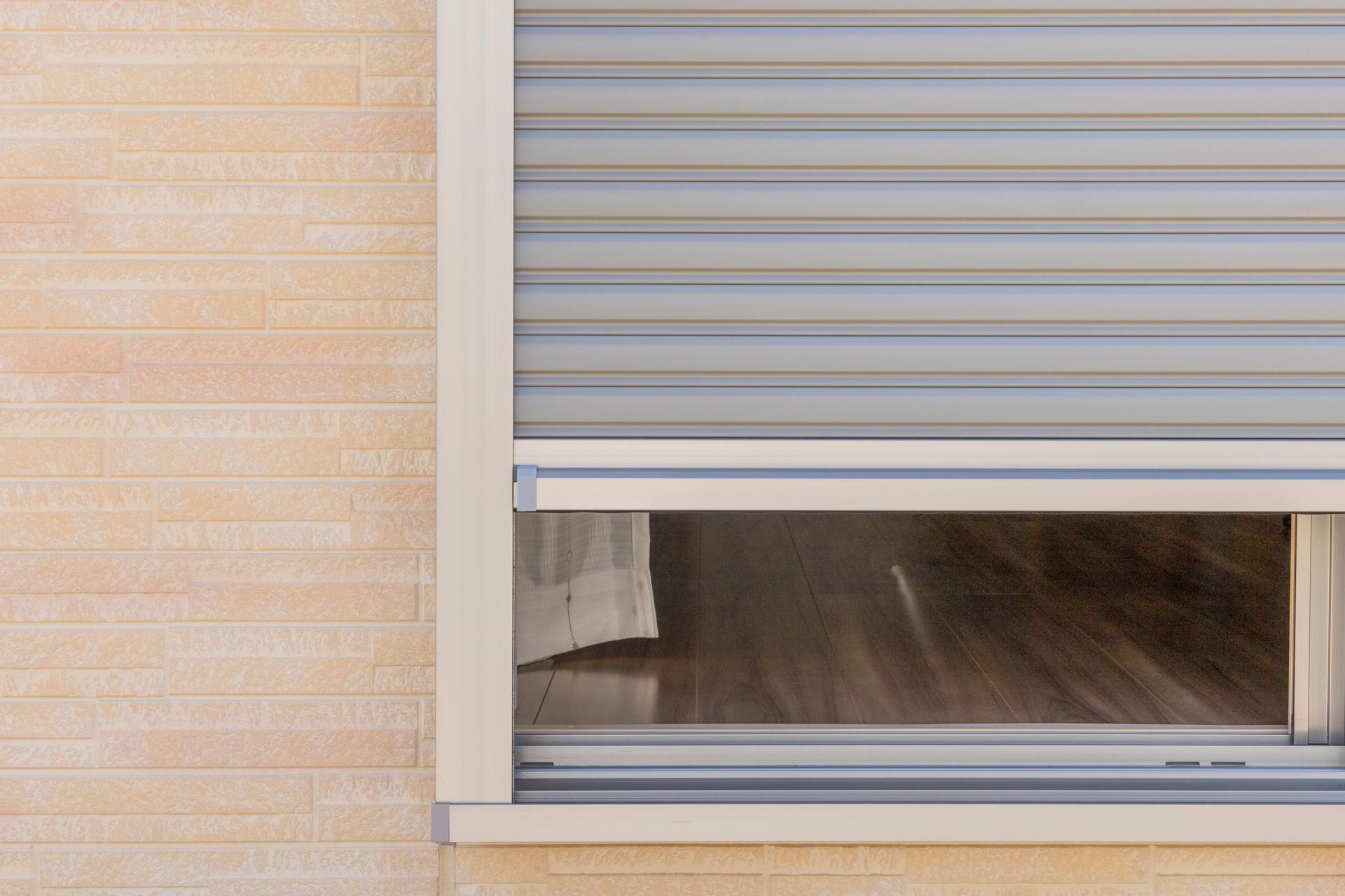 Exterior view of a window with partially lowered gray roller shutter on a beige brick wall.