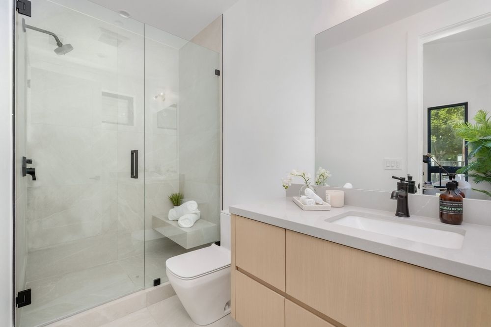 Modern bathroom with glass shower, floating vanity, toilet. Light wood tones, white surfaces, and black hardware.