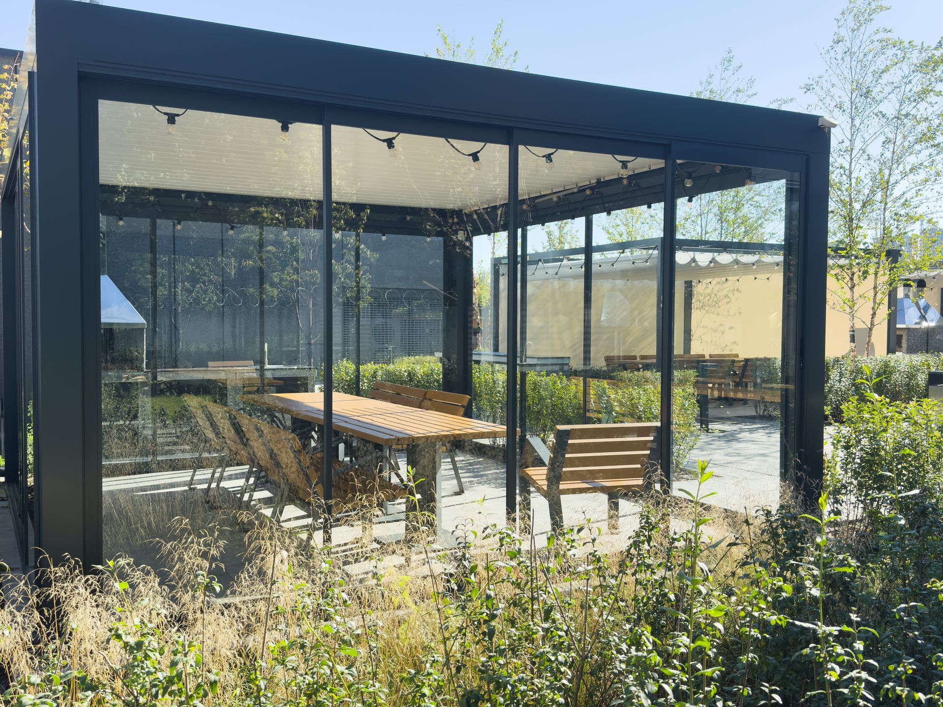 Outdoor dining enclosure with glass walls, wooden table and chairs, black frame, and a white canopy.