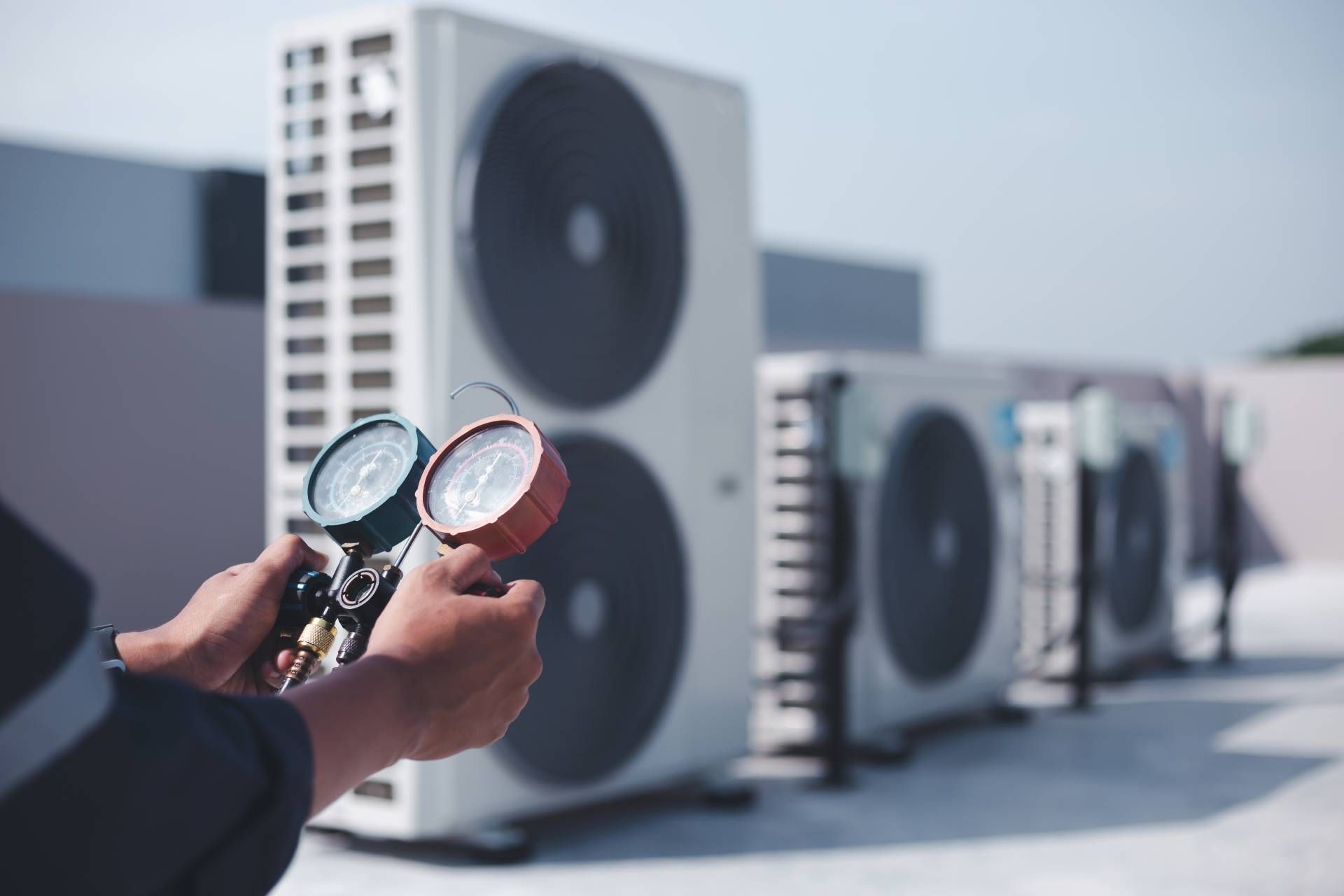 Hands holding pressure gauges in front of outdoor air conditioning units